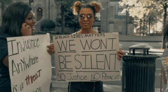 Two smiling black women stand holding handmade signs reading #Blacklives matter We Won't Be Silent NO Justice NO Peace and Injustice Anywhere is a Treat to Justice Everywhere