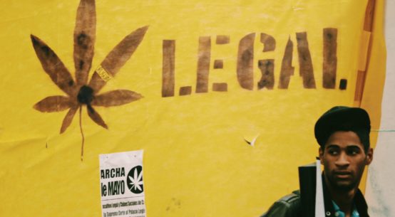 A young Black man with a backpack stands in front of a yellow banner behind him which reads Legal next to a cannabis leaf