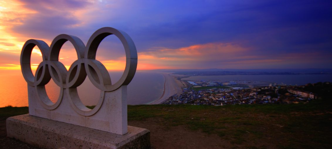 A statue of the five Olympic Rings sits above the ocean at sunset