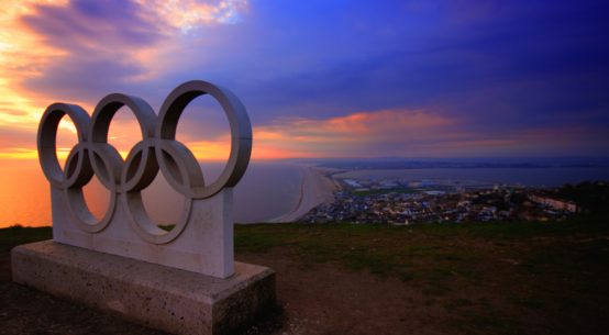 A statue of the five Olympic Rings sits above the ocean at sunset