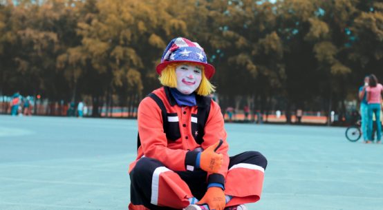 A sitting clown gives the thumbs up sign wearing a US flag hat